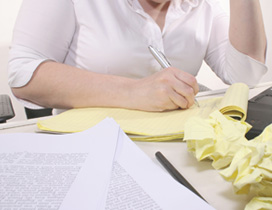 A close-up of women student at a desk; she actually is composing on a pad of paper. The work desk is covered in balls of crushed up paper. She is attempting to enhance the woman writing abilities.